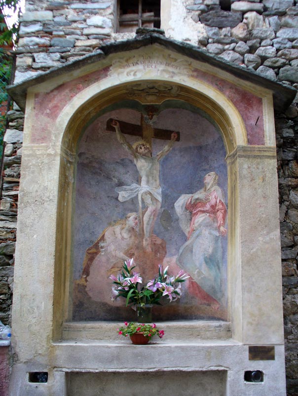 Cappelletta votiva con tetto in pietra raffigurante Gesù in croce e ai piedi di essa la Madonna e Maria Maddalena. Esterno affrescato in rosso scuro.
