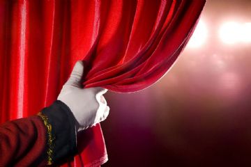 Una mano guantata di bianco scosta il sipario rosso di un teatro e si intravedono le luci della platea.