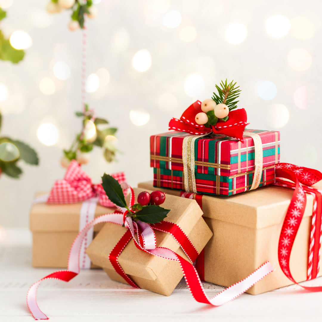 Sullo sfondo chiaro con luci natalizie quattro pacchi regalo, tre in scatole di carta colore beige e fiocchi rossi, una sovrapposta in scatola con motivo scozzese in rosso e verde e fiocco dorato con decorazione rossa.